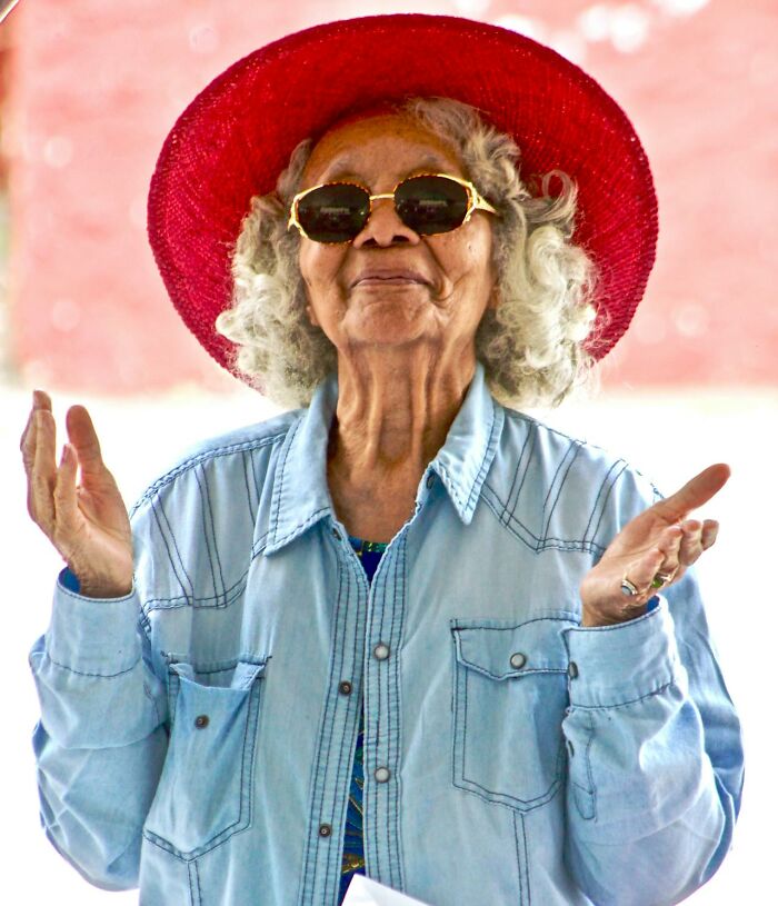 Smiling woman in sunglasses and red hat, gesturing with open hands, outdoors.