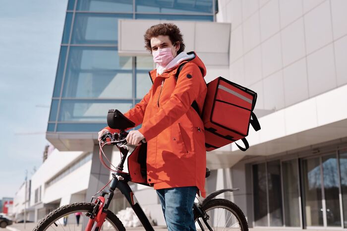 Delivery person wearing a mask with an insulated backpack, standing next to a bike in an urban area, highlighting restaurant food trends.