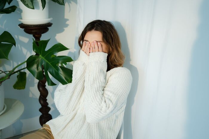 Woman embarrassed, covering face with hands in a cozy sweater, sitting by a plant and white curtain.