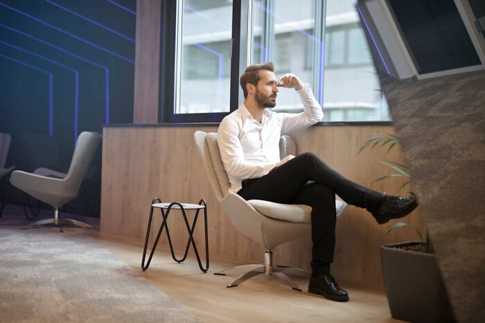 Man sitting in a modern lounge, pondering life abroad, dressed in a white shirt and black pants, near a large window.