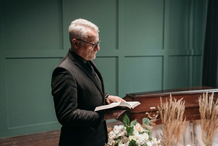 Man in black suit reading a book beside a coffin, symbolizing reasons to stop going to church.