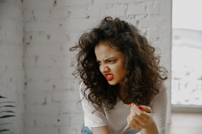 Woman with curly hair reacting with disgust, rolling her eyes and grimacing, indoors against a brick wall.