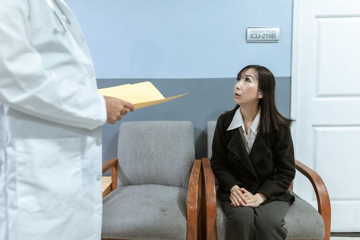 Woman sitting in hospital waiting room looking concerned while doctor stands nearby holding medical files, unfair treatment.