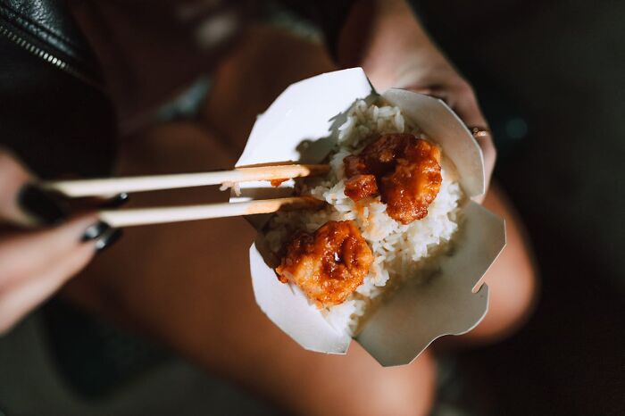 Chopsticks holding food from takeout box with rice in a potentially appalling dining setup.