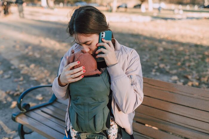 First responder making an urgent phone call while holding a baby in a carrier on a park bench outdoors.