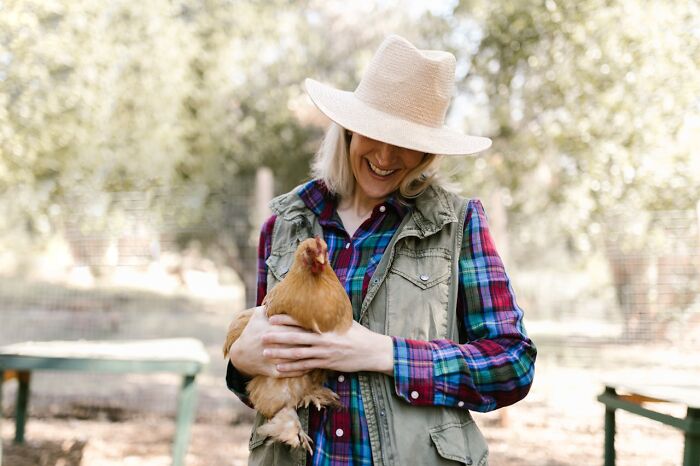 Person outdoors holding a chicken, wearing a hat and plaid shirt, with trees in the background, creating a relaxed atmosphere.