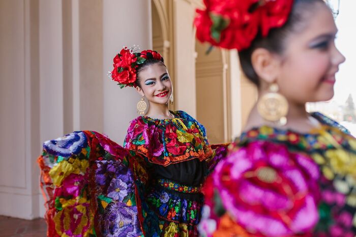 Two women in vibrant traditional dresses with floral patterns and red flowers in hair, showcasing rich cultural attire.