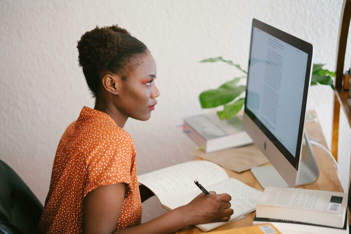 Woman sharing red pill stories, focused on writing notes while reading on a desktop computer in a home office.