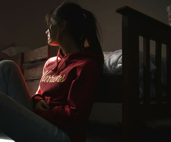 Young woman sitting on the floor in a dim room, appearing to be enduring unexplainable pain and deep in thought.
