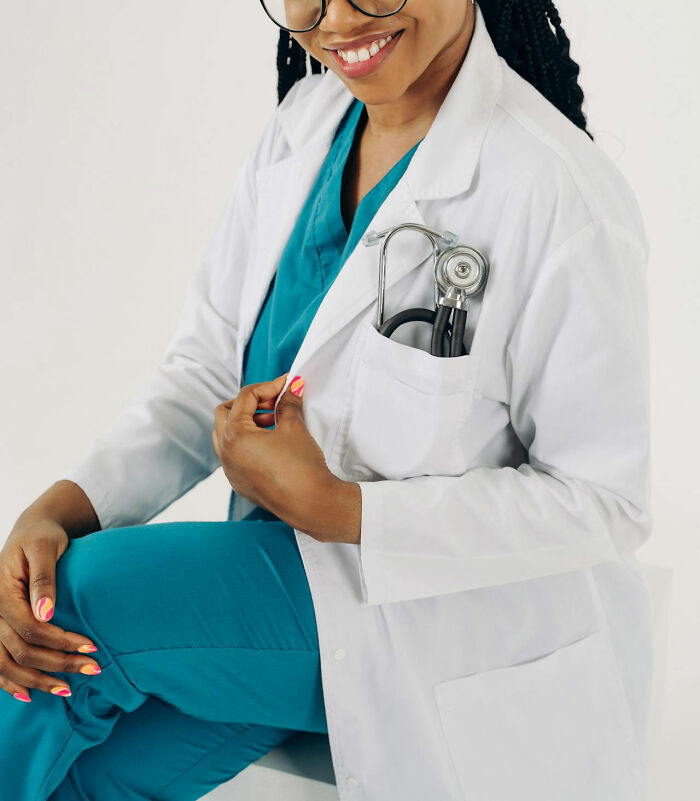American doctor in blue scrubs and white coat, smiling, with a stethoscope in pocket.