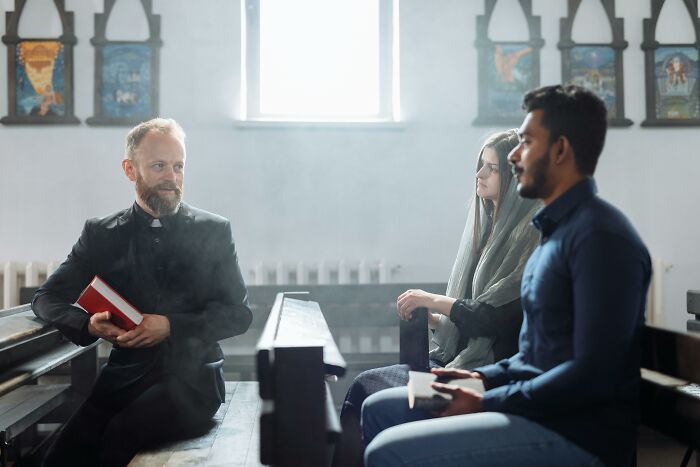 A priest holding a book talking to a man and woman in church pews, highlighting ghastly church experiences.