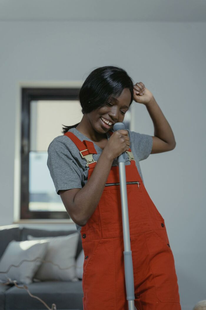 Person joyfully cleaning, wearing red overalls, holding a mop in a bright living room.