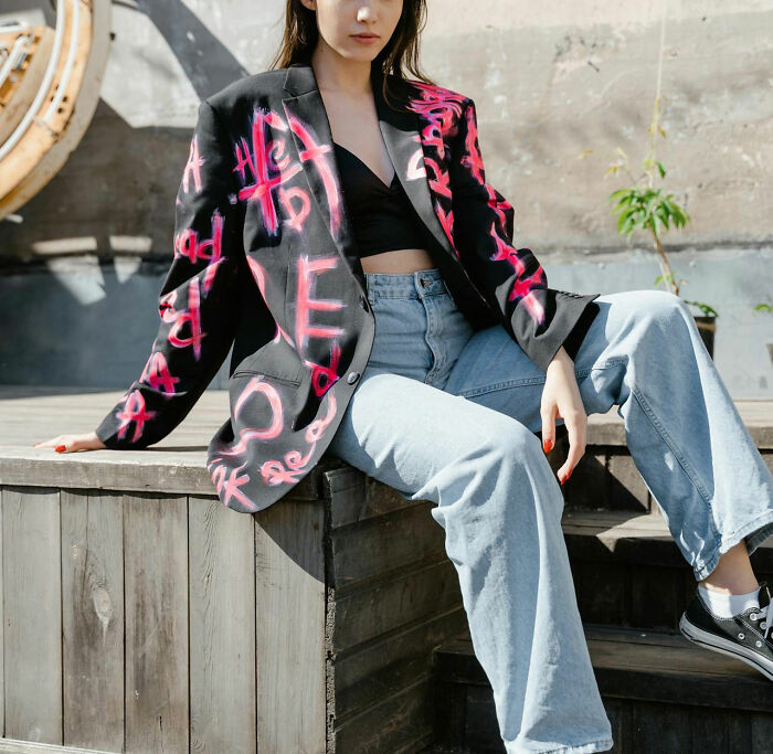 Woman wearing a graffiti-style blazer and jeans, seated outdoors.