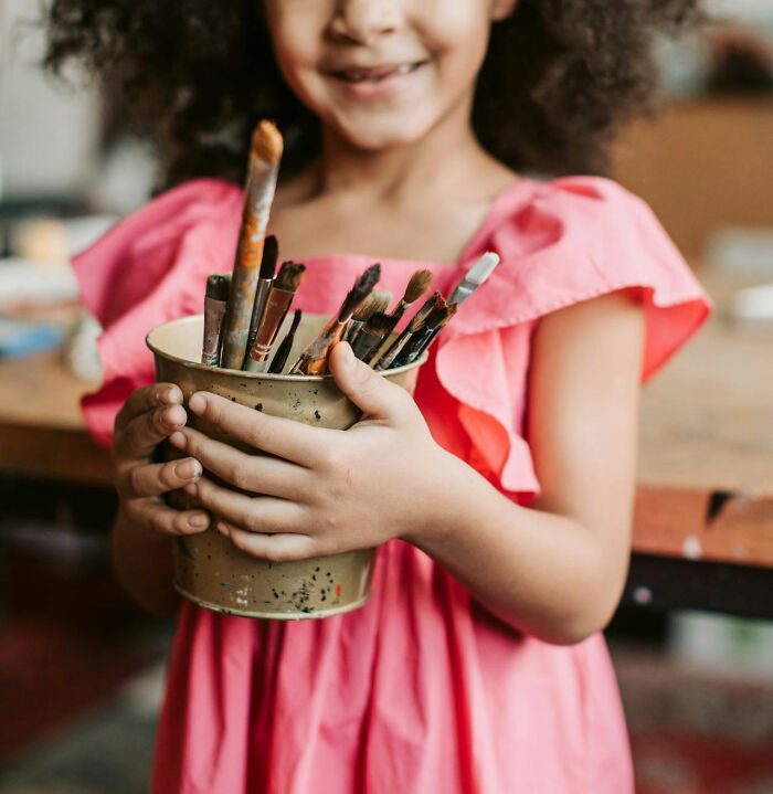 Child holding a cup of paintbrushes, wearing a pink dress, symbolizing creative mom sanity hacks.