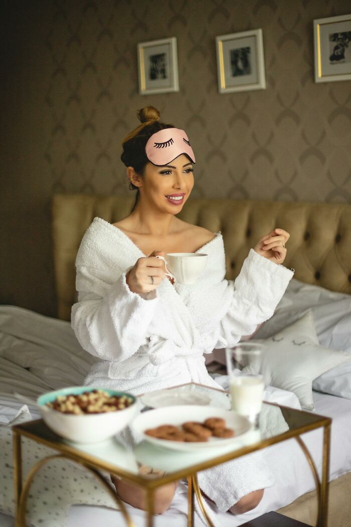 A woman in a bathrobe enjoys breakfast in bed, featuring cereal and cookies, embodying a creative mom sanity hack.