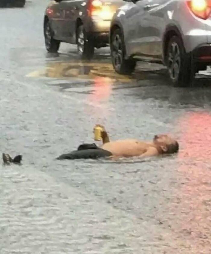 Man lying in flooded street holding a drink, creating a funny meme moment amidst traffic.
