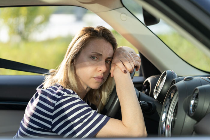 Woman in striped shirt looking concerned, resting on steering wheel, sunlight coming through car window.