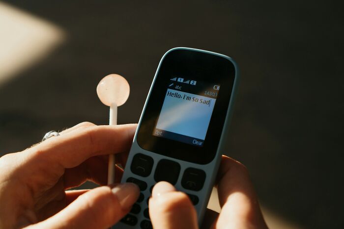 A hand holding an old mobile phone and a lollipop, with a sad text message displayed on the screen.