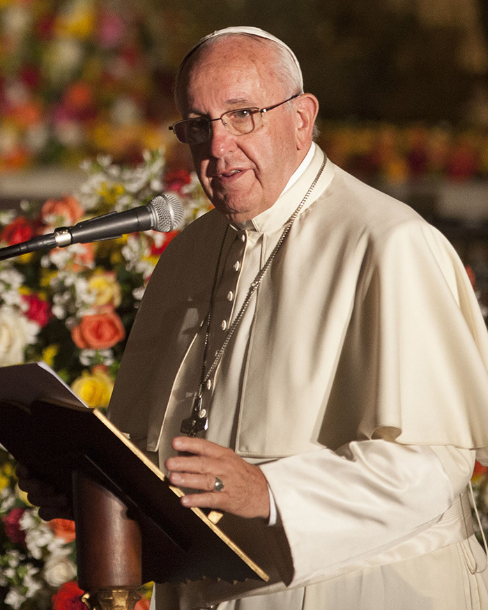 Pope Francis speaking at an event, wearing traditional papal attire, with floral decor in the background.