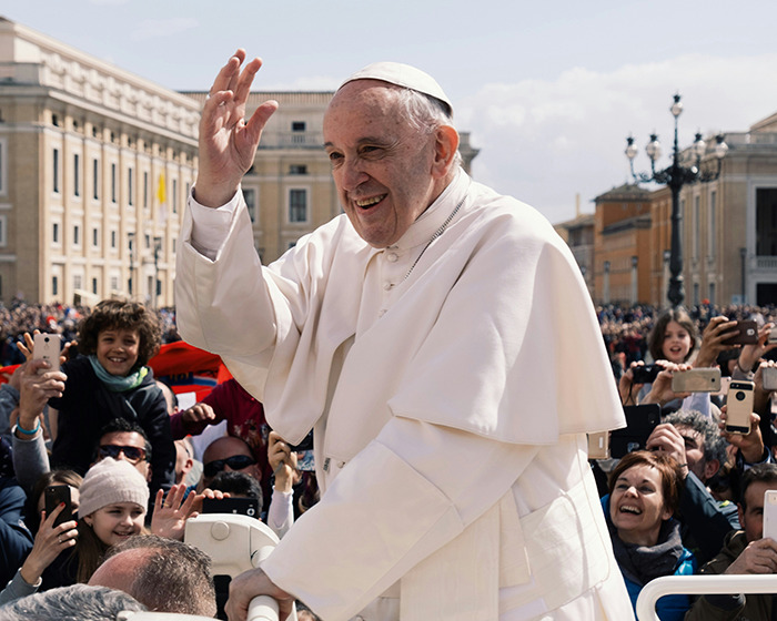 Man in white robe greeting crowd in outdoor setting, surrounded by smiling people and cameras.