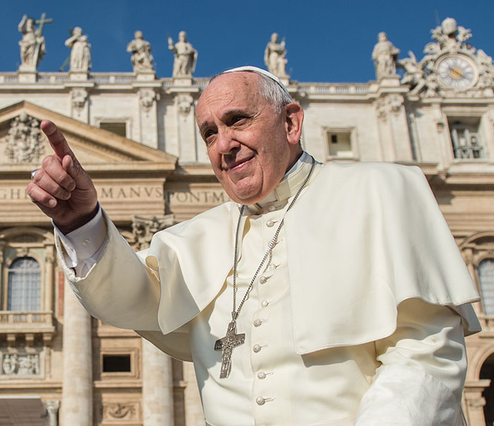 The Pope smiling and gesturing outdoors near the Vatican in formal attire.