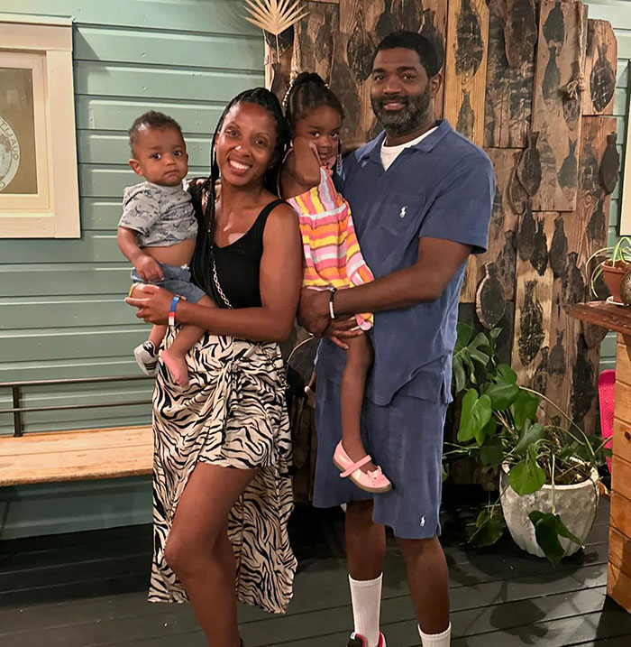 A family of four smiling inside a wooden-themed room, with two adults holding kids, during a casual photo shoot.