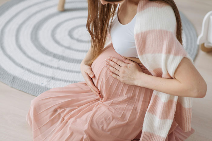 Pregnant woman in a pink skirt sitting on the floor, gently touching her belly.