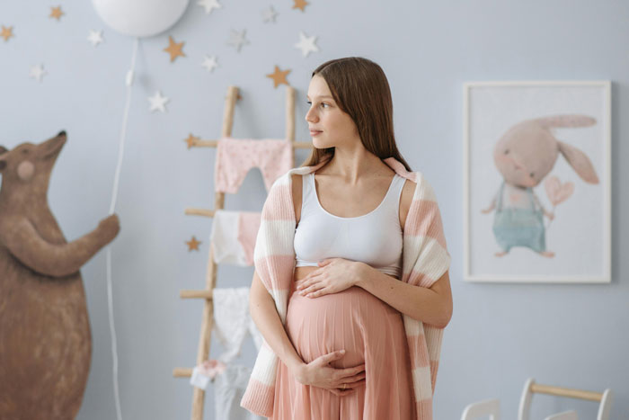 Pregnant woman in a pastel-themed nursery, gently holding her belly, surrounded by whimsical decor.