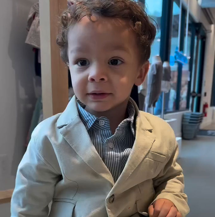 Toddler in a light-colored suit smiles indoors.