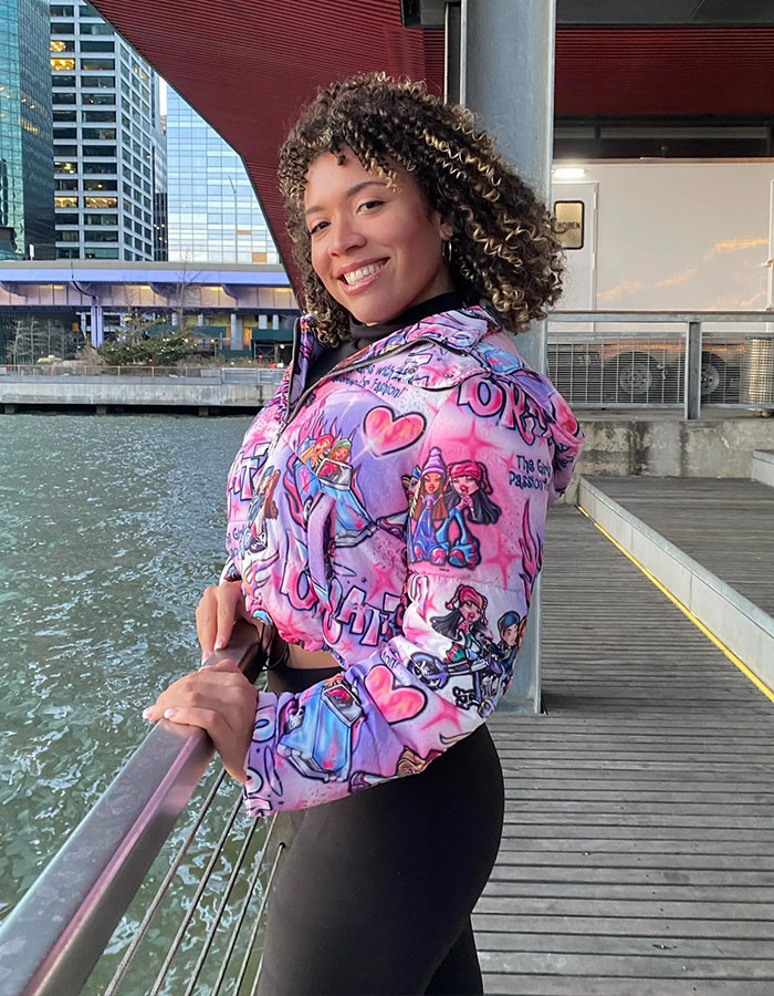 Smiling woman in colorful jacket by waterfront, standing outside near modern buildings.