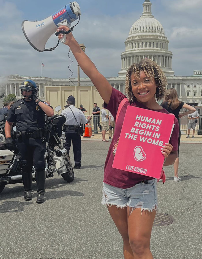 Pro-life activist holds megaphone and sign reading "Human Rights Begin in the Womb" near the Capitol building.