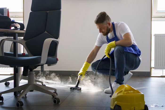 Man using steam cleaner on office floor for quality life improvement, wearing gloves and apron.