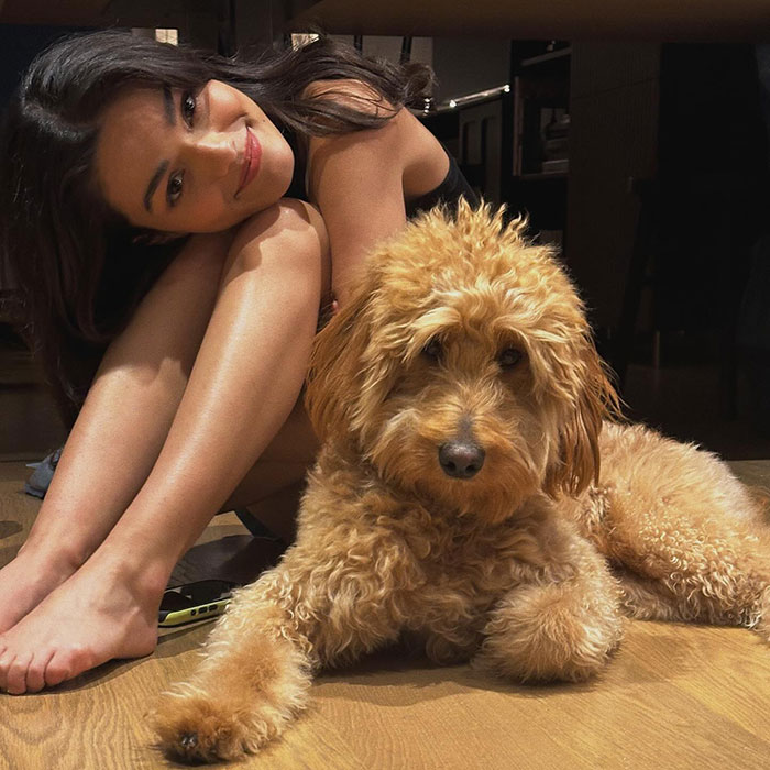 Woman sitting beside a fluffy dog on a wooden floor, looking at the camera.