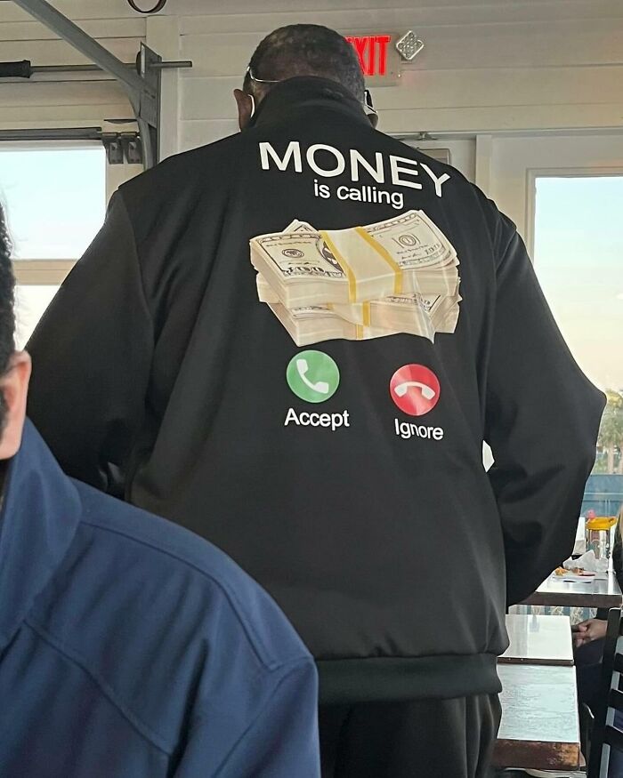 Man wearing a jacket with funny image of cash and text "Money is calling, accept or ignore" in a humorous setting.