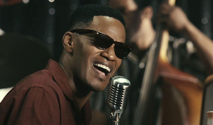 Actor wearing sunglasses singing into a vintage microphone, showcasing dedication in challenging roles on stage performance.