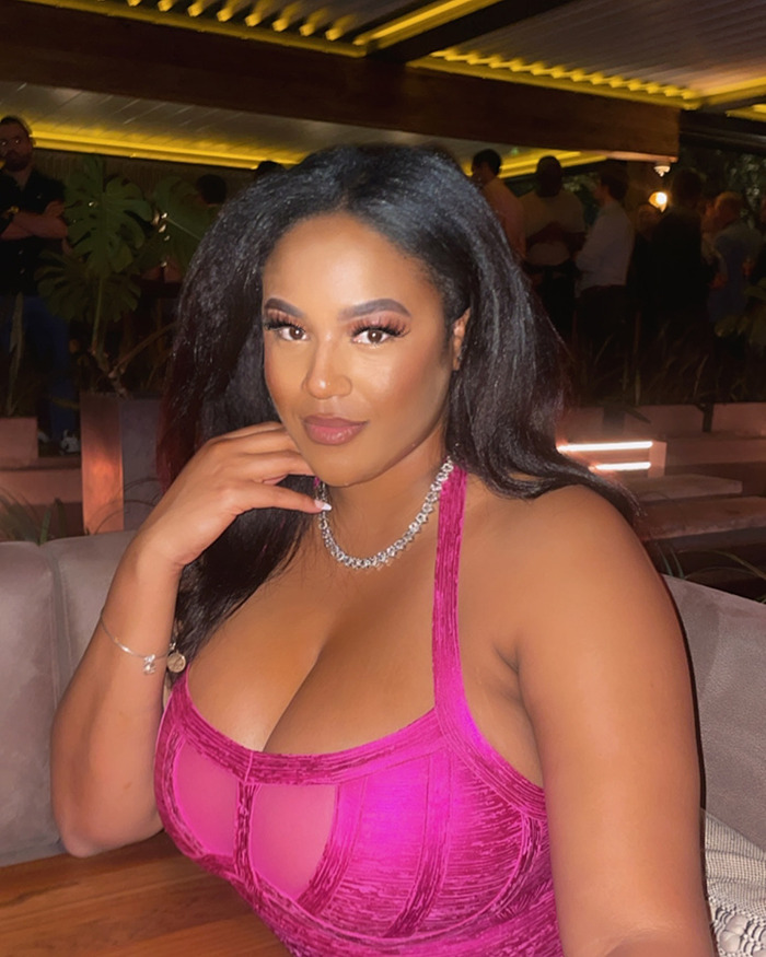 A woman in a pink top seated in a dimly lit restaurant, involved in a viral video story about her hair being braided by a stranger.