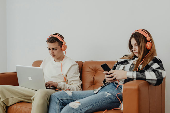 Two teens with headphones, one using a laptop and the other a phone, embodying entitled attitude and parental challenges.