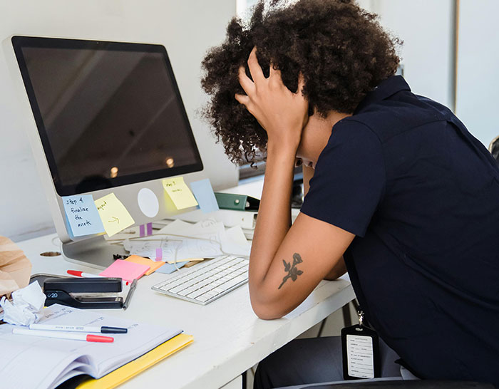Person stressed at work, sitting at a cluttered desk with computer and notes, reflecting recession impact.