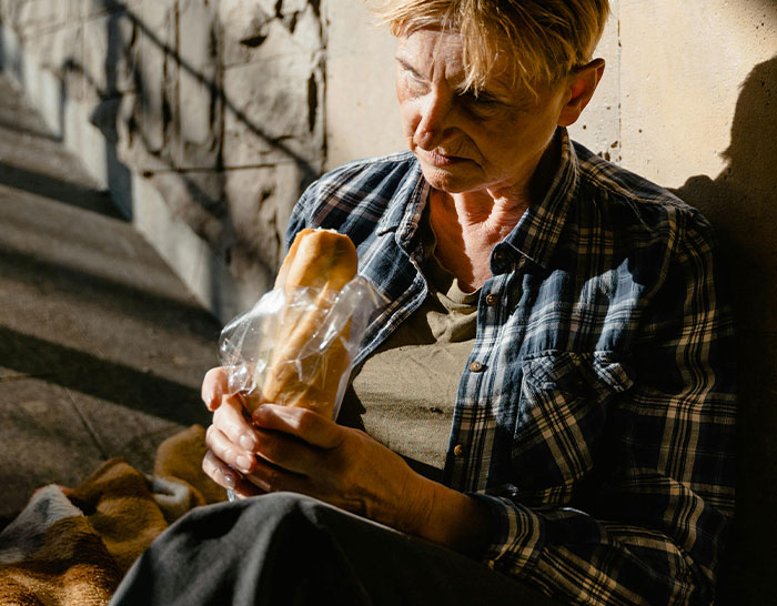 Person in a flannel shirt sitting outdoors, holding a packaged baguette, illustrating recession's impact on daily life.