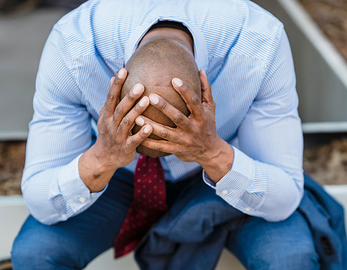 Man in a striped shirt and red tie sits with head in hands, symbolizing stress during a recession.