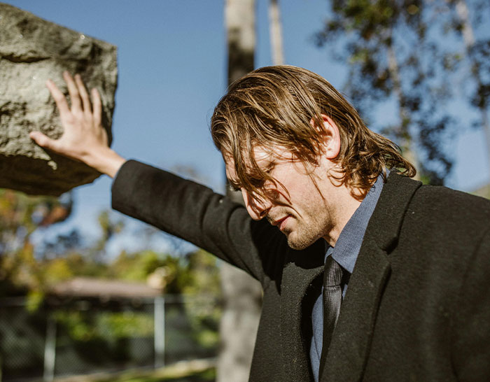 Man in a suit leaning on a large rock, pondering recession experiences outdoors.