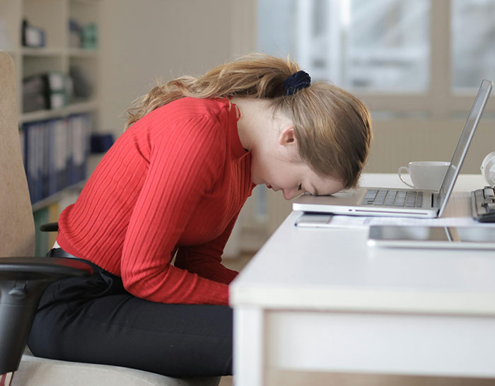 Woman in red shirt, head on desk, showing stress during a recession while using a laptop in an office setting.