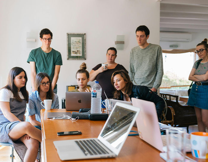 People gathered, attentively discussing a recession, with laptops on a large table in a casual meeting room.