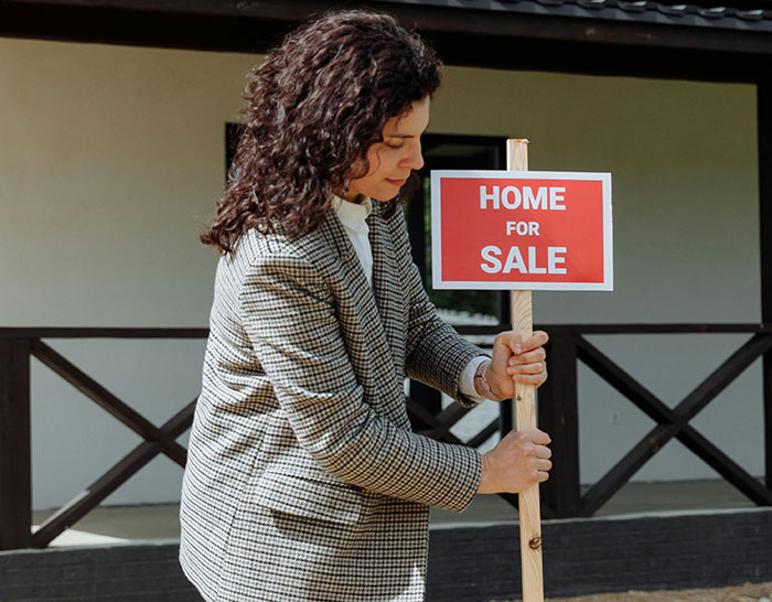 Woman placing a "Home for Sale" sign outside a house, symbolizing recession challenges in real estate.