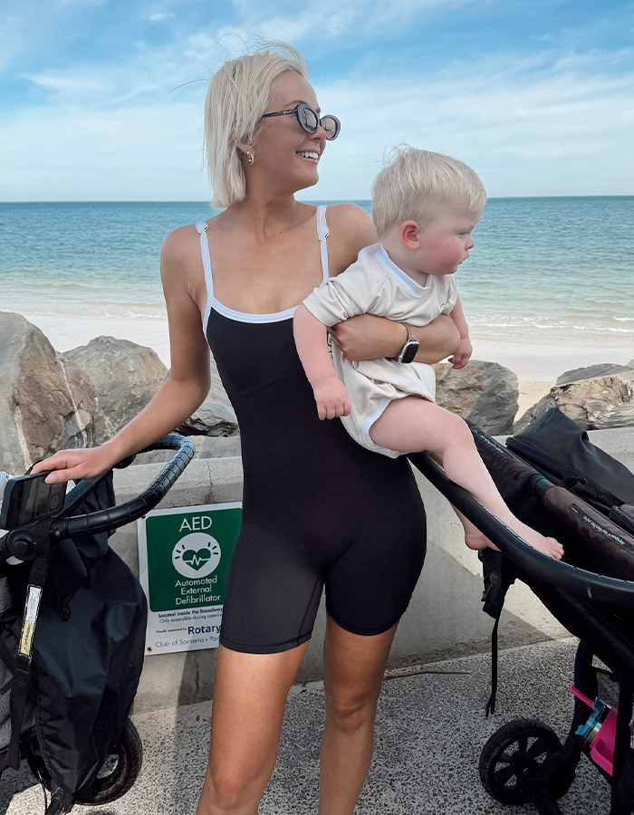 Woman holding a toddler near the beach, surrounded by strollers, with a cheerful expression.