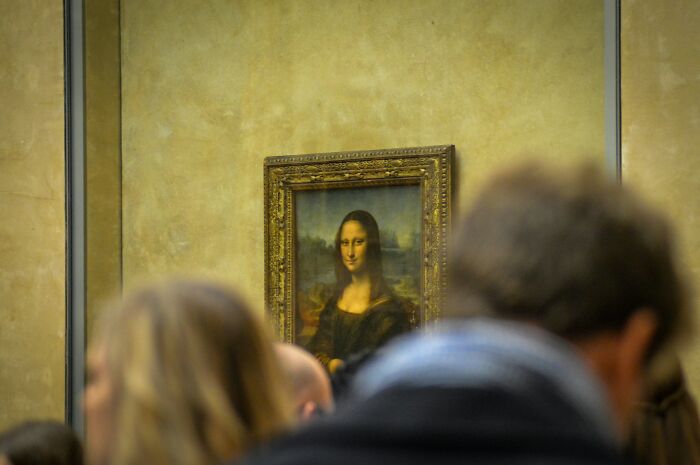 Tourists crowd around the Mona Lisa, capturing a disappointing FOMO experience at the Louvre Museum.