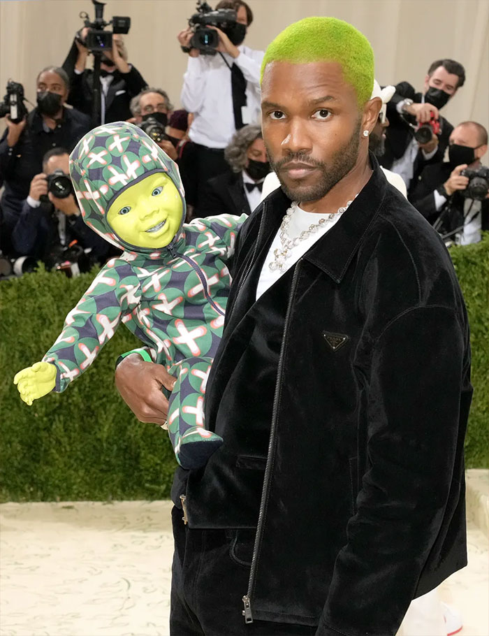 Frank Ocean with neon green hair holding a green-faced doll dressed in a bold outfit at the Met Gala fashion event.