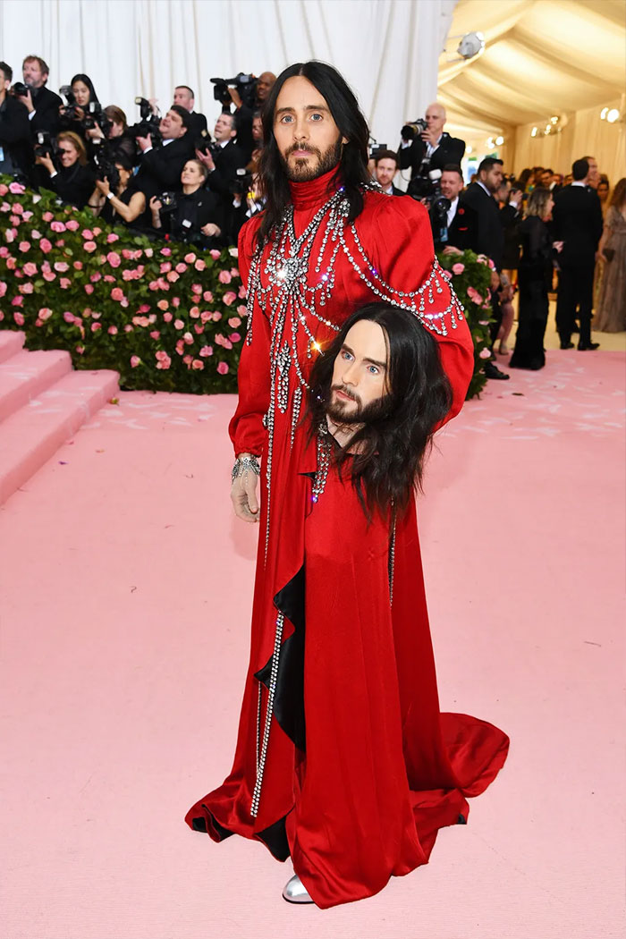 Celebrity in an elaborate red Met Gala outfit holding a realistic replica of his own head on a pink carpet event.