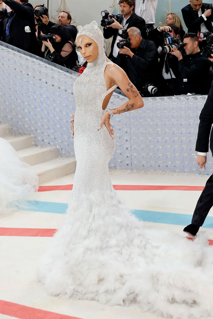 Celebrity in a sparkling white gown with a dramatic feathered train posing amid photographers at the Met Gala event.