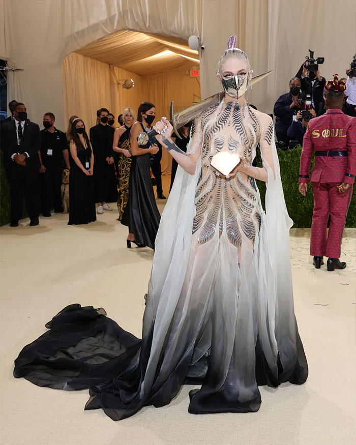 Celebrity in avant-garde outfit with metallic accessories and long ombré gown at Met Gala, showcasing bold fashion choices.